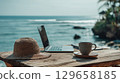 Remote work setup with laptop, coffee, and hat on a wooden table by the ocean 129658185