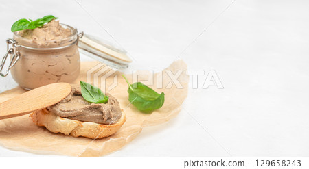 Jar of creamy pate with basil, served on toasted sesame bread slice with a wooden spreader, on parchment paper light background. copy space 129658243