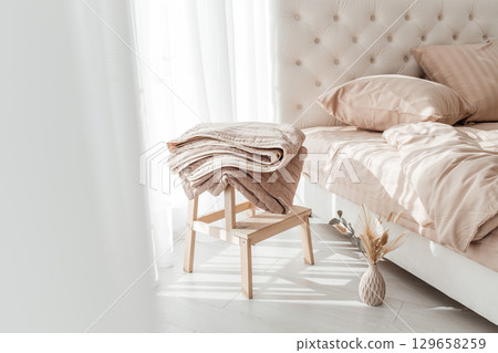 Cozy folded beige blanket on wooden stool next to a neatly made bed in morning sunlight 129658259