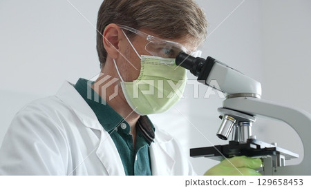 Masked adult scientist analyzing microscope slides, wearing green protective gear while conducting detailed medical research in sterile laboratory environment. Medicine, healthcare and science concept 129658453