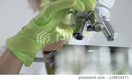 Close up of green gloved hands of a scientist adjusting a microscope in a laboratory, emphasizing the significance of scientific research and analysis. Medicine, healthcare and science concept 129658471