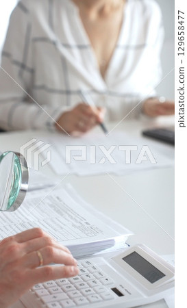 Two accountants are working together, using a magnifying glass and a calculator, to audit financial reports in their brightly lit office. Audit and taxes in business 129658477