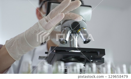 Scientist wearing white gloves adjusting microscope in laboratory setting, conducting scientific research surrounded by test tubes. Medicine, healthcare and science concept 129658479
