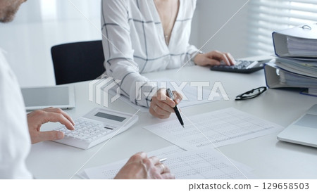 Professional women collaborating, reviewing financial reports using calculator, laptop, discussing workplace strategy with team. Audit and taxes in business Professional women collaborating, reviewing financial reports using calculator, laptop, discussing workplace strategy with team. Audit and taxes in business 129658503
