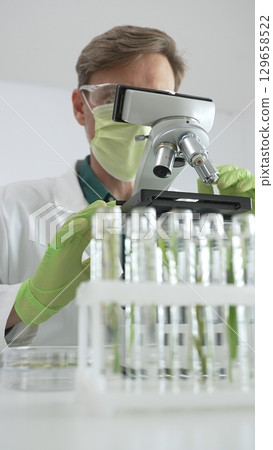 Male scientific researcher in green protective gear analyzing specimens through a microscope in a sterile laboratory environment. Medicine and science concept 129658522