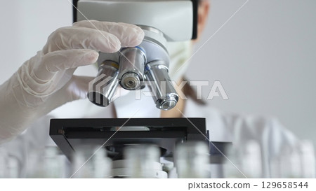Scientist wearing white gloves adjusting microscope in laboratory setting, conducting scientific research surrounded by test tubes. Medicine, healthcare and science concept Scientist wearing white gloves adjusting microscope in laboratory setting, conducting scientific research surrounded by test tubes. Medicine, healthcare and science concept 129658544
