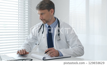 A male doctor in a lab coat with a stethoscope sits at a table in a medical office and uses a laptop while taking notes on a clipboard for patient care and treatment 129658629