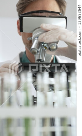 Man scientific researcher in protective gear analyzing specimens through a microscope in a sterile laboratory environment. Medicine, health care and science concept Man scientific researcher in protective gear analyzing specimens through a microscope in a sterile laboratory environment. Medicine, health care and science concept 129658644