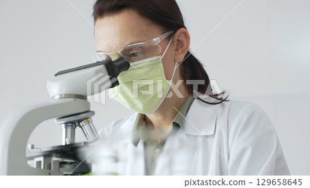 Female professional scientist examining specimen, placing slide under microscope while wearing green protective gear in laboratory workspace. Medicine, health care and science concept 129658645