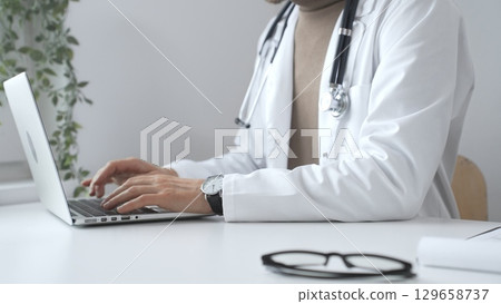 Male doctor wearing a white coat and stethoscope is typing on a laptop in a bright medical office, showcasing the intersection of healthcare and technology, close up. Medicine and health care concept 129658737