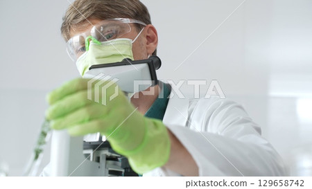 Adult male scientist wearing green mask, gloves and goggles using microscope and analyzing sample in test tube in a laboratory, developing new drugs and vaccines. Medicine and science concept 129658742