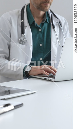 Doctor man wearing a white coat over green shirt, and stethoscope is typing on a laptop in a bright medical office, showcasing the intersection of healthcare and technology. Medicine and health care 129658829