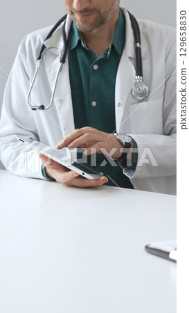 Doctor man wearing lab coat over green shirt, and stethoscope, is using digital tablet in modern medical office, browsing patient information or conducting online consultation. Medicine and health 129658830