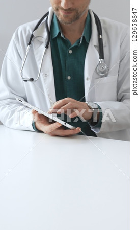 Doctor man wearing lab coat over green shirt, and stethoscope, is using digital tablet in modern medical office, browsing patient information or conducting online consultation. Medicine and health Doctor man wearing lab coat over green shirt, and stethoscope, is using digital tablet in modern medical office, browsing patient information or conducting online consultation. Medicine and health 129658847
