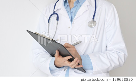 Unknown female doctor wearing stethoscope and white coat over blue blouse is standing and writing on a black clipboard, representing medical expertise and patient care. Medicine concept 129658850