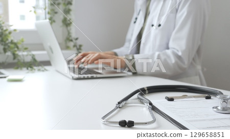 Stethoscope and medical records are on the desk opposite the doctor is typing on a laptop with a in a medical office, suggesting online healthcare or telemedicine services. Medicine concept 129658851