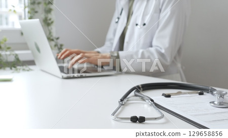 Stethoscope and medical records are on the desk opposite the doctor is typing on a laptop with a in a medical office, suggesting online healthcare or telemedicine services. Medicine concept 129658856