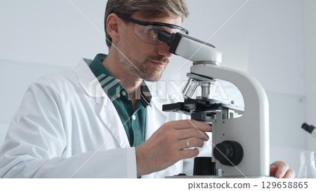 Adult male scientist wearing lab coat and protective glasses using microscope, doing scientific research in modern laboratory. Medicine, healthcare and science concept Adult male scientist wearing lab coat and protective glasses using microscope, doing scientific research in modern laboratory. Medicine, healthcare and science concept 129658865