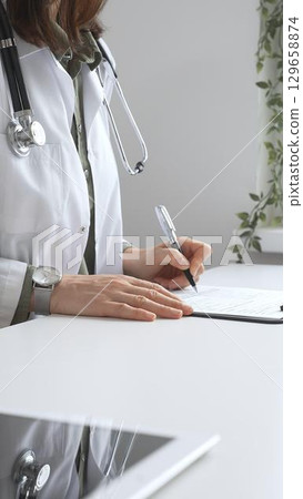 Professional female doctor wearing white lab coat and stethoscope documenting patient information on clipboard near laptop and digital tablet in clinic workspace. Medicine and health care concept 129658874