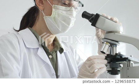 Female scientist examining microscope slide, wearing white protective gear while conducting medical research in laboratory environment. Medicine, health care and science concept 129658875