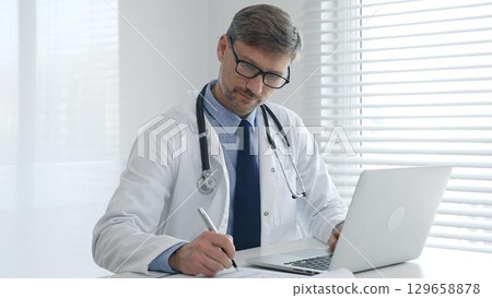 A male doctor in glasses and a lab coat with a stethoscope is typing on a laptop keyboard while sitting at a table in a bright medical office 129658878