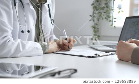 Doctor woman listening to male patient health concerns and writing a prescription on a clipboard and office table during a consultation in a bright, modern clinic, close up. Medicine and health care 129658889