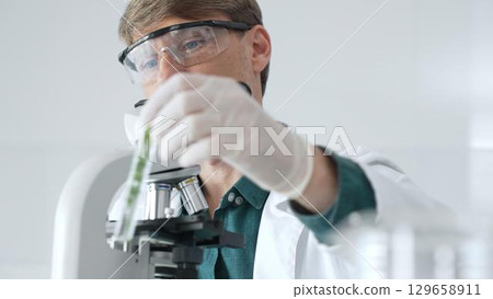 Adult male scientist in a lab coat and gloves examining a plant sample in a test tube, with a microscope set up in the foreground. Medicine, healthcare and science concept 129658911