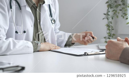 Doctor woman listening to male patient health concerns and writing a prescription on a clipboard and office table during a consultation in modern clinic, close up. Medicine concept 129658925