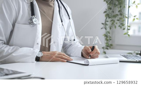 Male medical professional working at desk, writing prescription, wearing white lab coat and stethoscope in clinical workspace. Medicine and health care Male medical professional working at desk, writing prescription, wearing white lab coat and stethoscope in clinical workspace. Medicine and health care 129658932