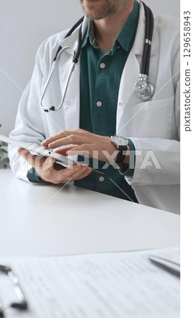 Doctor man wearing lab coat over green shirt, and stethoscope, is using digital tablet in modern medical office, browsing patient information during online consultation. Medicine and health care Doctor man wearing lab coat over green shirt, and stethoscope, is using digital tablet in modern medical office, browsing patient information during online consultation. Medicine and health care 129658943
