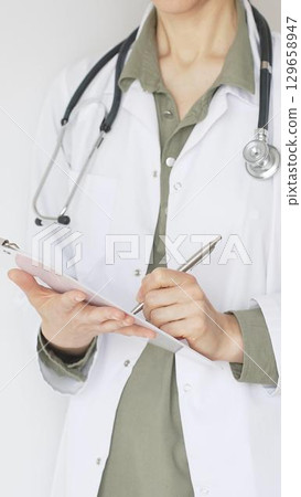 Female doctor, wearing a stethoscope and a medical coat over a green blouse, is standing and writing on a white clipboard, representing medical expertise and patient care. Medicine concept. Female doctor, wearing a stethoscope and a medical coat over a green blouse, is standing and writing on a white clipboard, representing medical expertise and patient care. Medicine concept. 129658947