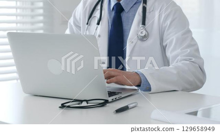 Male doctor in lab coat and stethoscope sitting at desk typing on laptop, holding glasses and pen nearby in medical office. Medicine and healthcare concept 129658949