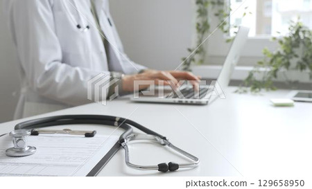 Stethoscope and medical records are on the desk opposite the doctor is typing on a laptop with a in a medical office, suggesting online healthcare or telemedicine services. Medicine concept Stethoscope and medical records are on the desk opposite the doctor is typing on a laptop with a in a medical office, suggesting online healthcare or telemedicine services. Medicine concept 129658950