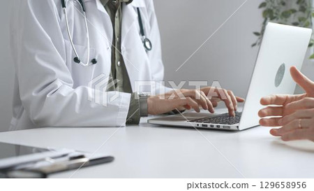 Doctor wearing lab coat and stethoscope typing on laptop keyboard and explaining something to patient during a medical consultation in hospital office. Medicine and health care concept 129658956