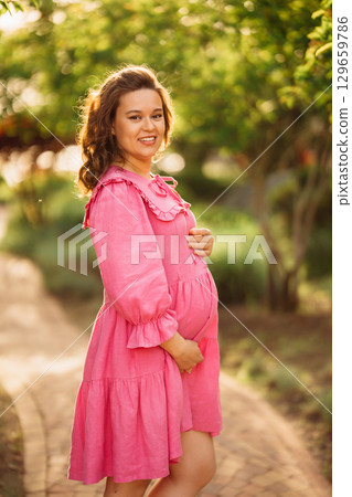 Beautiful and happy pregnant young woman in pink dress on a walk in the park 129659786