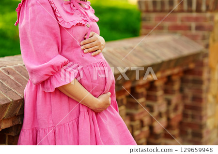 pregnant woman in pink dress hugging belly by the stone fence in the park pregnant woman in pink dress hugging belly by the stone fence in the park 129659948