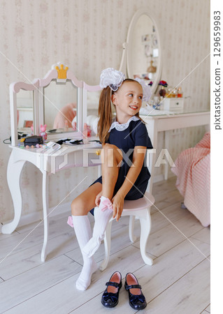 A little girl in a school uniform puts on knee socks at a dressing table.  129659983