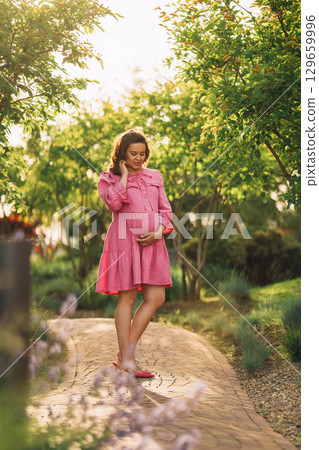 Beautiful and happy pregnant young woman in pink dress on the path in the park Beautiful and happy pregnant young woman in pink dress on the path in the park 129659996