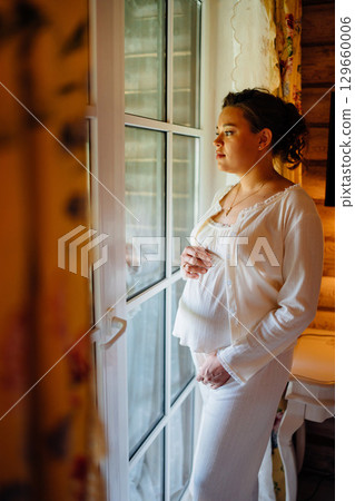 Calm and beautiful young pregnant woman in white pajamas by the window.  129660006