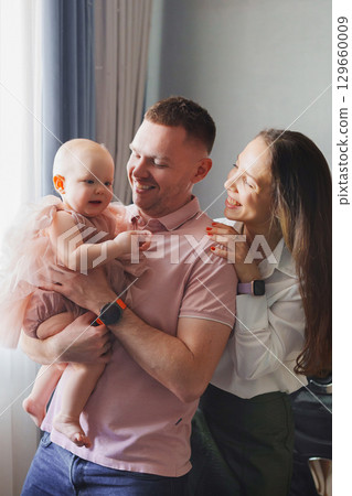 Happy and gentle parents hold baby daughter in pink dress in their arms. Happy and gentle parents hold baby daughter in pink dress in their arms. 129660009
