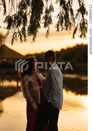 man and pregnant woman in red dress at sunset on the riverbank 129660012