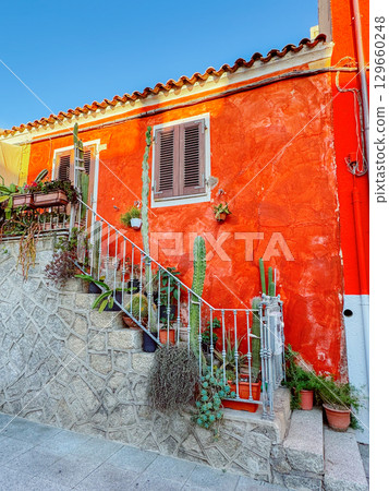 Charming red house in Sardinia with rustic stone stairs, potted cacti and Mediterranean plants. Perfect scene for travel, architecture, lifestyle, home decor, tourism, and real estate projects. 129660248
