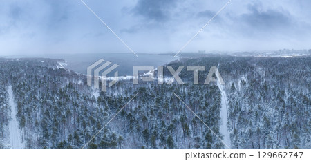 Snow-covered forest on lake shore with the first ice at sunset and the city on horizon, aerial view Snow-covered forest on lake shore with the first ice at sunset and the city on horizon, aerial view 129662747
