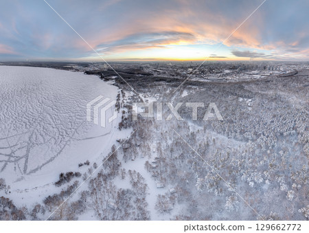 Snow-covered forest on lake shore with ice at sunset and the city on horizon, auerial view 129662772
