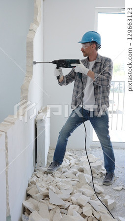Man construction worker wearing grey checked shirt and blue hardhat is demolishing a wall with a hammer drill, creating construction debris on the surrounding floor surface. Renovation and repair 129663123