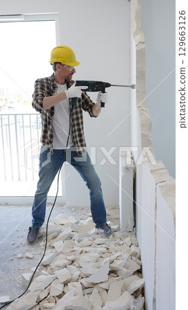 Construction worker demolishing concrete wall with rotary hammer, wearing safety gear, generating rubble and dust during extensive home renovation process 129663126