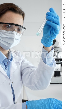 Scientific researcher holding test tube, analyzing blue liquid while wearing protective gear during medical experiment in sterile laboratory setting. Medicine, health care and science concept 129663134