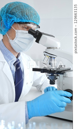 Senior male scientist wearing a protective mask, cap, and gloves, examining samples under a microscope in a modern laboratory. Medicine, healthcare and science concept 129663135