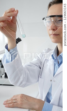 Scientific researcher holding test tube, analyzing blue liquid while wearing protective gear during medical experiment in sterile laboratory setting. Medicine, health care and science concept 129663143