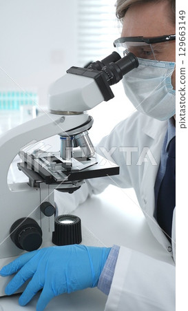 Man scientist wearing protective googles and blue gloves, is using microscope analyzing samples in modern laboratory doing scientific research. Medicine and science concept Man scientist wearing protective googles and blue gloves, is using microscope analyzing samples in modern laboratory doing scientific research. Medicine and science concept 129663149